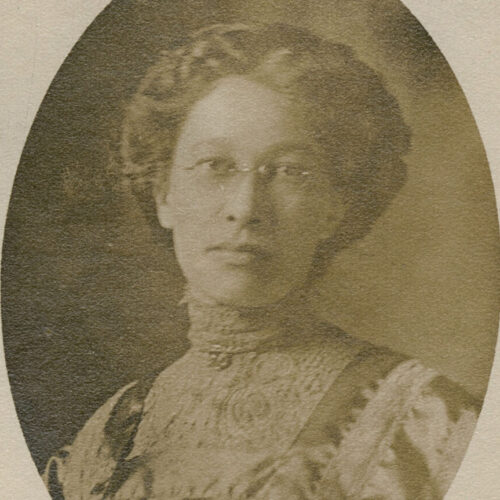 She is standing front- facing. The photo is from her mid chest up. Her hair is in “up bun” style. She is wearing thin frame glasses. The photo is faded black, grey and white. She is wearing a white flowery top button up to her neck.