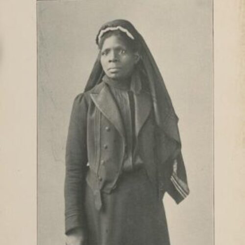 She is standing and wearing a long black skirt, Black blouse covered with a black jacket with black buttons. She’s also wearing a long black headscarf with white stripes on the bottom. The picture is black against a grey faded background