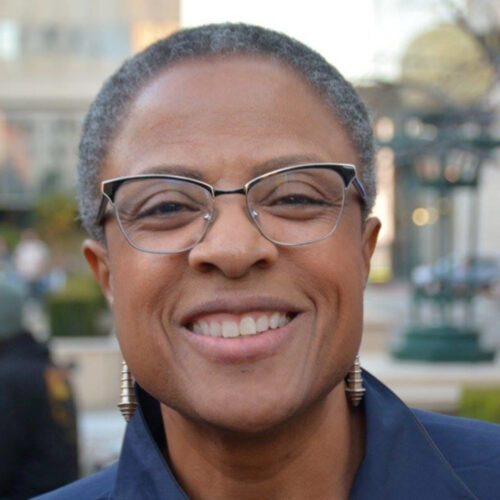 Yvette Flunder smiles with black rimmed cat-eye-glasses. Her hair is styled as a short black and grey afro. She is wearing dangling earrings and a collared top.