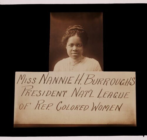 A young Nannie Helen Burroughs stands behind a sign that reads “Miss Nannie H. Burroughs President Nat’l League of Rep. Colored Women .” She is wearing a bloused with a collar around her neck. She has a part down the middle of her head and her hair is pulled up in a top bun.