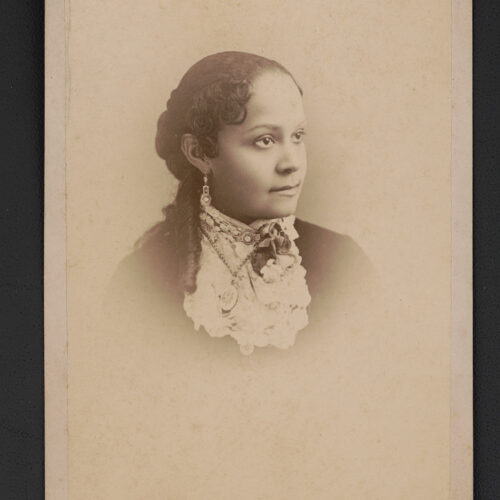 Black and white faded photo of her from mid chest up. She’s looking off to the side. She is wearing a white ruffled blouse over a black top. The blouse is buttoned to the top with a pendant. She also has hanging earrings.