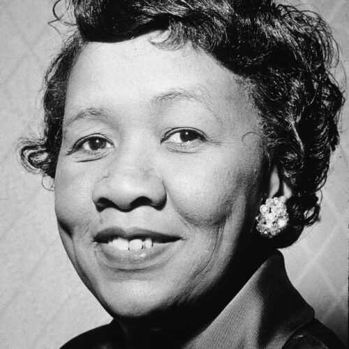 Dorothy Height is smiling. She is wearing a dark satin top with a beaded necklace and rhinestone earrings. Her hair is short and curled.