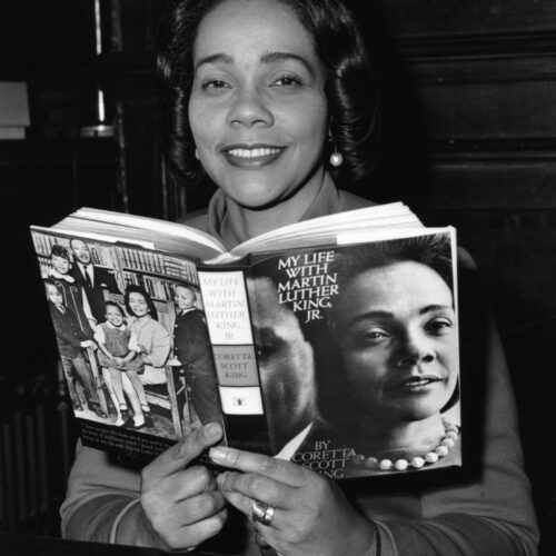 Coretta Scott King is seated holding an open copy of her book (My life with Martin Luther King Jr.). The book jacket features a picture of the King family on the back and a portrait of her with pearls and a right profile of Dr. King’s face on the front. She is smiling with dangling pearl earrings her hair in a bouffant of curls that frame her face.