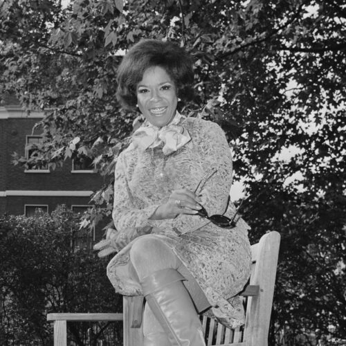 Della Reese: A black and white photo of a young Della Reese is seated with her left leg crossed over her right on the arm of a chair. The setting is outdoors with grass and leaves in the foreground and a large maple tree and trimmed bushes in the background. She is smiling wearing a dress with a scarf tied around her neck in a bow. She has pearl earrings in a short curled bob bouffant and knee-high boots on.