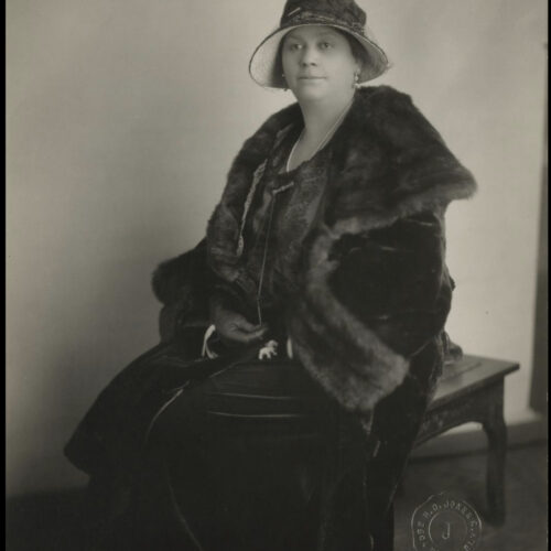 Lethia Cousins Fleming is seated on a bench profiled to the right. She is wearing a large fur over her shoulders that drapes down on top of her velvet dress. She is wearing a pair of dangling earrings and beaded hat with a hat pin in the front.