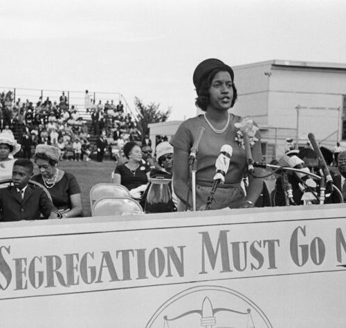 Myrlie Evers Williams stands at an outdoor podium with several microphones of different sizes. A large sign in front of the podium reads “Segregation Must Go Now." Immediately behind her are people seated with suits, dresses, and hats. Further in the background are people seated in bleachers. Myrlie is captured mid sentence and is wearing a short-sleeved shirt and skirt, as well as a pearl necklace and button hat. Her hair cascades the her face in a curled side bang.
