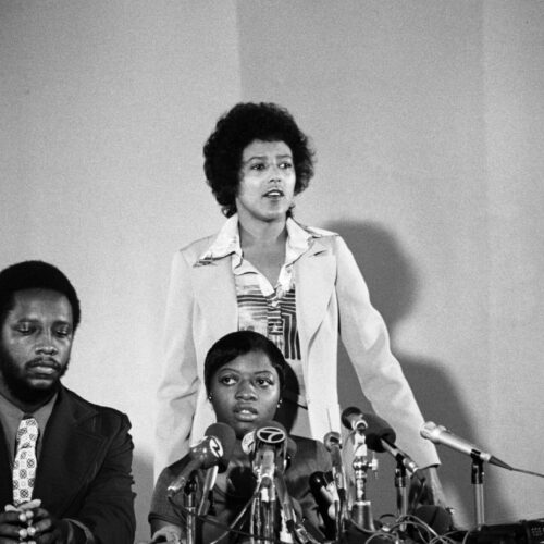 Elaine Brown stands behind her client Joanne Little at a press conference. She is wearing a light colored suit and patterned blouse, while her hair is styled in a soft curled afro with a slight bang.