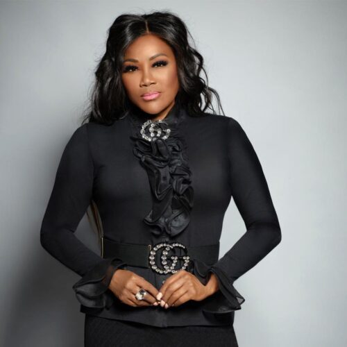 Headshot of Juanita Bynum from the waist up. Her hair is styled in large soft curls down her back. She is wearing a black dress with ruffles down the center and a matching collar and belt bedazzled broach.