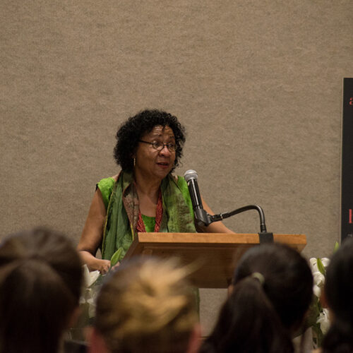 bell hooks stands at a podium with a microphone looking toward a crowd of women. She is wearing flowing scarf, thin framed glasses, and her hair is styled in soft natural curls. In the background is a large poster of the front cover of her book, All About Love.