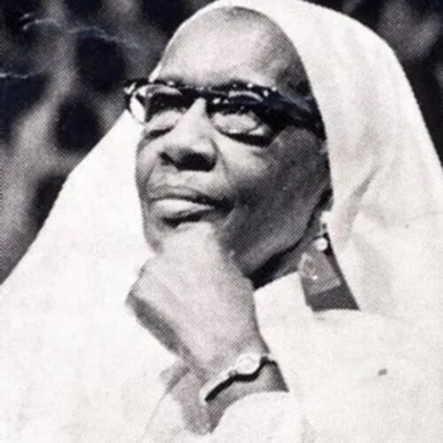 Clara Muhammad is wearing glasses, a watch, and a veil. She is looking toward the left with her chin on her hand. Photo is in black and white.