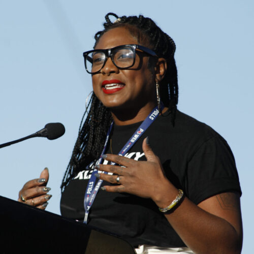 A colored photo of her at a podium speaking. She has on a black top. She is wearing glasses.