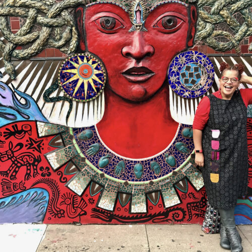 Faith Adiele stands to the right of a colorful outdoor mural of a deity. She is wearing a patterned black dress on top of a red long sleeve shirt. Her natural hair is pulled into two ponytails. She is wearing red and black rimmed glasses and has a joyful smile on her face.