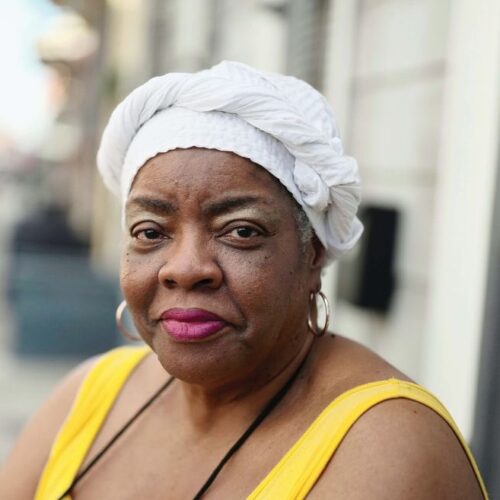 Portrait of Luisah Teish wearing a white head scarf, gold hoop earrings, and a yellow sleeveless top.