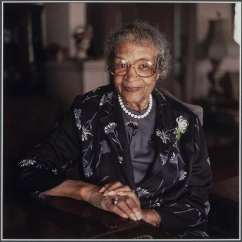 Frances Albrier has gray hair and glasses. She is smiling and wearing a dark purple suit with soft white flowers and a white necklace, sitting with hands folded.