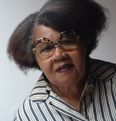 Jamaica Kincaid is facing the camera leaning left. Kincaid's hair is parted on the left side. The photo is from the mid-chest. Kincaid is wearing large tortoise shell glasses and a black and white vertically striped shirt.
