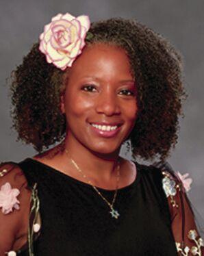 Photo of Dr. Tarece Johnson looking forward and smiling. She has a white and pink flower in her curly shoulder length hair. She is wearing a black dress that has pink and green flowers on the sheer sleeves. Dr. Johnson has a Star of David pendant hanging from her necklace.