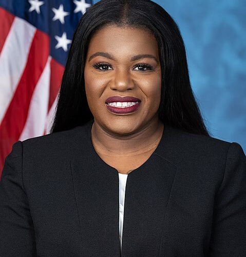 Cori Bush faces front and is wearing a black jacket with no collar. A small opening at the top of the jacket shows a light blue and white blouse. She has a slight smile. The photo is from her mid chest up. A portion of the U.S. flag is visible over and behind her right shoulder. The rest of the background is blue.