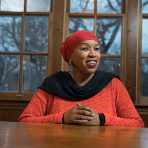 Photo of Debra Majeed facing forward with windows and trees in the background. She is seated with her hands crossed resting on a brown table. She is dressed in a modest red top with a black scarf loosely draping her neck. Her copper colored danglings earrings frame her face and compliment her complexion. Majeed is wearing a red hijab and smiling as she looks at the camera.