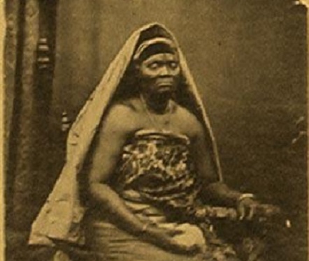 Efunroye Tinubu is posed sitting in a chair. She is wearing an African wrap dress with bangles on her wrists. Her head is covered by a long piece of cloth that cascades to her waist. One hand is positioned on the arm of the chair and the other is in her lap. The image is in black and white.