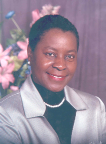Dr. Jacquelyn Grant is posed in the portrait facing forward. She has on a silver jacket, black blouse, pearls, and small stud earrings. She has short hair and is wearing red lipstick. The photo is from the chest up and in the background are pink flowers.