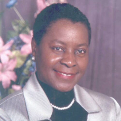 Dr. Jacquelyn Grant is posed in the portrait facing forward. She has on a silver jacket, black blouse, pearls, and small stud earrings. She has short hair and is wearing red lipstick. The photo is from the chest up and in the background are pink flowers.