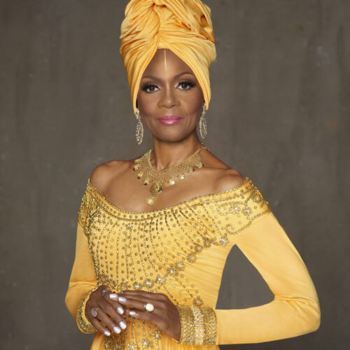 Queen Afua is posed, facing front with a slight smile. They are wearing a tall head-wrap and a long-sleeved dress with dropped shoulders. The dress is ornamented around the neckline. The head-wrap and dress are yellow gold. Queen Afua is wearing gold earrings and an ornate gold neclace. The photo is cropped near the bottom of the torso showing Queen Afua's bent arms and touching hands with white nail polish, a ring with a white stone on the left hand, and two gold rings on the right hand.