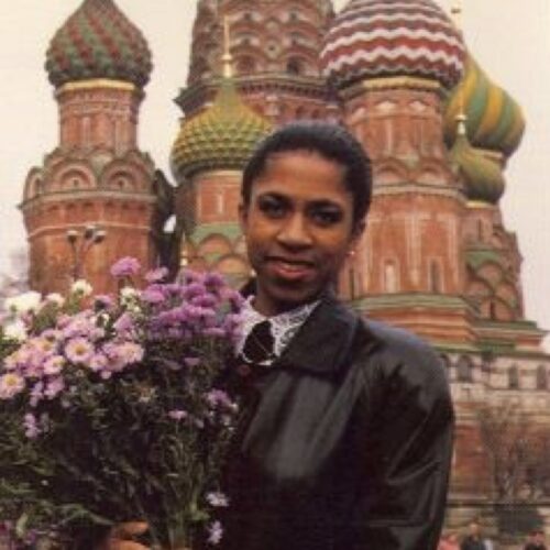 Yelena Abdulaevna Khanga stands holding a bouquet of pink and white daisies in the crook of her right arm. Fingers of her left and right hands are interlaced at the bottom of the bouquet. She is smiling and wearing a black leather jacket. The photo is from her mid abdomen up. The domes of St. Basil’s Cathedral on Red Square, Russia, are in the background.