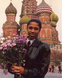 Yelena Abdulaevna Khanga stands holding a bouquet of pink and white daisies in the crook of her right arm. Fingers of her left and right hands are interlaced at the bottom of the bouquet. She is smiling and wearing a black leather jacket. The photo is from her mid abdomen up. The domes of St. Basil’s Cathedral on Red Square, Russia, are in the background.