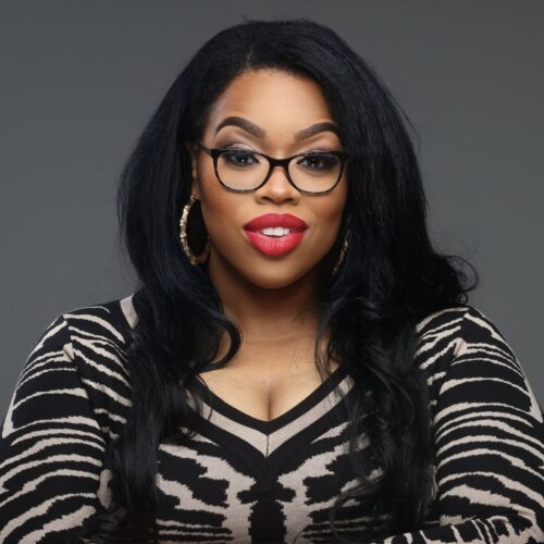 Photo of Candice Benbow in front of a solid grey background. She is wearing a zebra print blouse, large gold hooped earrings, red lipstick, black glasses, and long straight black hair with loose curls at the ends.