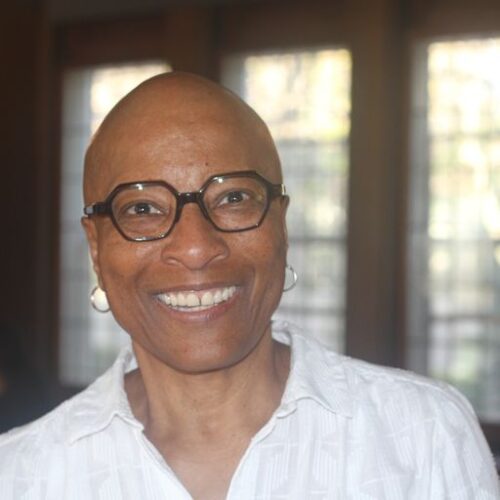 Photo of a Black woman facing the camera and smiling. She has on a white blouse and small hoop earings. She has black framed glasses and a shaven head.