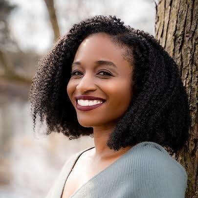 Headshot of Khristi Adams leaning against a tree. She is wearing a grey sweater, smiling with marron lipstick, and curly shoulder-length hair.