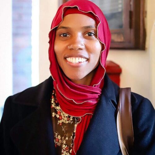 Headshot of Dr.Jamillah Karim smiling. She is wearing a red Hijab and navy blue winter coat