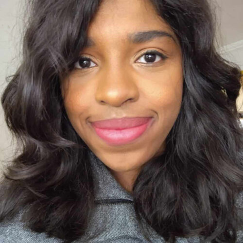 Photo of Maayan Zik. She is a black woman with brown, shoulder-length, wavy hair that is parted to the side, smiling with pink lipstick.
