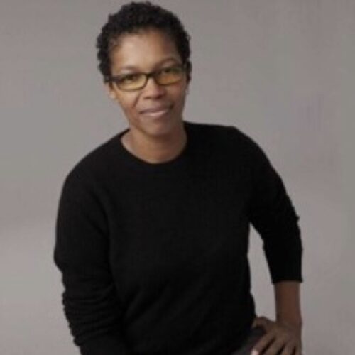 Angel Kyodo Williams is standing in front of a plain gray background, smiling, wearing a black sweater and glasses, with her left hand propped on her side.