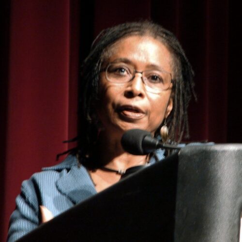 Alice Walker has shoulder-length dreadlocks, glasses, earrings, blue suit, standing at a podium with microphone.