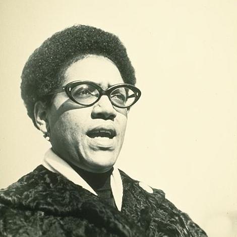 Audre Lorde is wearing glasses and looking toward right. Her hair is in a short afro. She is wearing a dark patterned coat with a white scarf tucked in and a dark turtleneck. Photo is in 2-tone sepia.