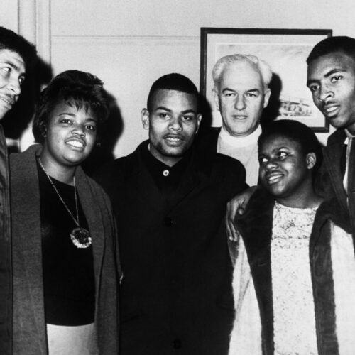 Bernice Johnson is standing 5th from the right among 6 people. She is looking left and smiling. She has on a lace shirt with an open white coat with dark trim. Also pictured are Charles Neblett, Ruth Harris, Cardell Reagan, Rev. Eddy, and Carver Neblett. Photo in black and white.