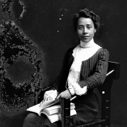 Anna Julia Cooper has wavy hair pulled back. She is wearing a ruffled high-neck white shirt and pleated jacket with a skirt. She is sitting holding an open book, gazing straight ahead. Photo in black and white.