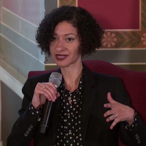 Chanda Prescod-Weinstein has short curly black hair and dark rose lipstick. She is holding a microphone, and wearing a black jacket with a black and white polka dot shirt, and a gold ring on each hand.