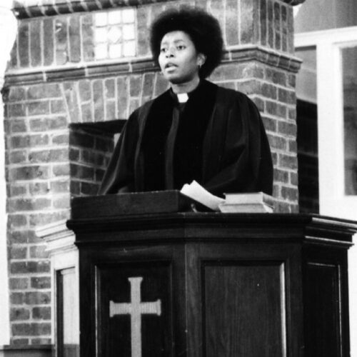 Katie Cannon is wearing a black robe with a clerical collar, standing behind a pulpit with 2 books on it and a cross on the front. Her hair is in a medium Afro, she is wearing small hoop earrings and she is speaking. The background is brick.