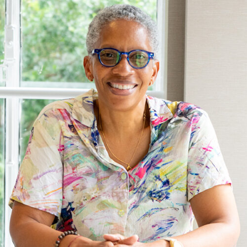 Pamela Ayo Yetunde is smiling, her hair short and gray, she is wearing glasses, gold earrings, and a multicolored short-sleeved shirt. She is sitting with a window and greenery behind her.