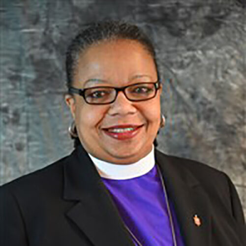 Robin Dease is smiling, wearing glasses, red lipstick. gold earrings, a clerical collar with a purple shirt, black jacket, and a gold pin. The background is gray.