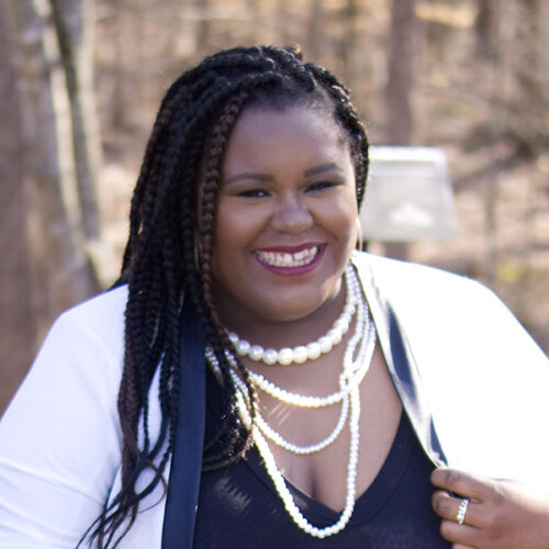 D. Danyelle Thomas has long braided hair. She is smiling, wearing 4 strands of white beads, a black V-neck shirt and a white jacket. She is holding her jacket lapel with her left hand and wearing on a ring on that hand. The woods are behind her.