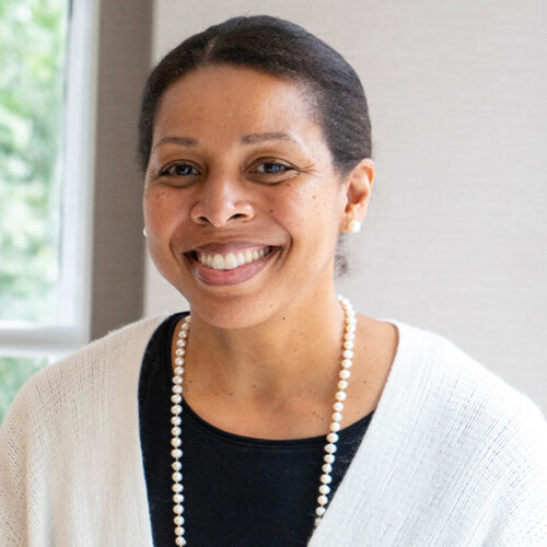 Karen Jackson-Weaver is smiling, wearing a pearl necklace, earrings, a black shirt with a white sweater, and her hair pulled back. There is a a partial window in the background.