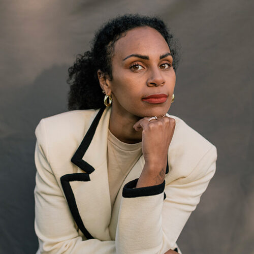 Rachel Ricketts is wearing gold earrings, red lipstick, her hair pulled back in a pony tail, and a cream colored suit with a brown lapel outline. She is sitting leaning forward on her elbow with her left hand under her chin.