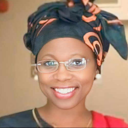 Sindile Dlamini Mathebula is wearing an orange and dark gray headscarf tied fashionably, white earrings, glasses, and pink lipstick, with a gray and red top and a gold necklace. She is smiling.