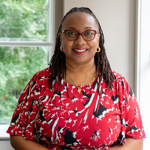 Tawnicia Rowan is smiling, wearing dark red glasses, red lipstick, gold earrings, shoulder length braids, a gold necklace and pendant, and a red dress with white and black abstract designs and short sleeves.