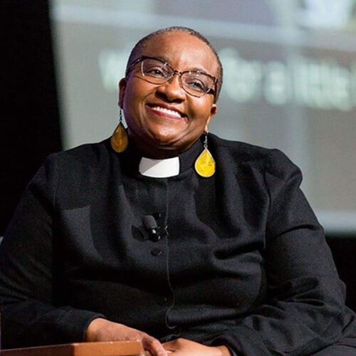 Nontombi Naomi Tutu is smiling, wearing glasses, a clerical collar, gold dangling earrings, sitting and leaning to her right.