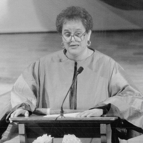 Delores Williams is speaking, standing behind a podium. She is wearing a robe, glasses, and earrings. The photo is in black and white.