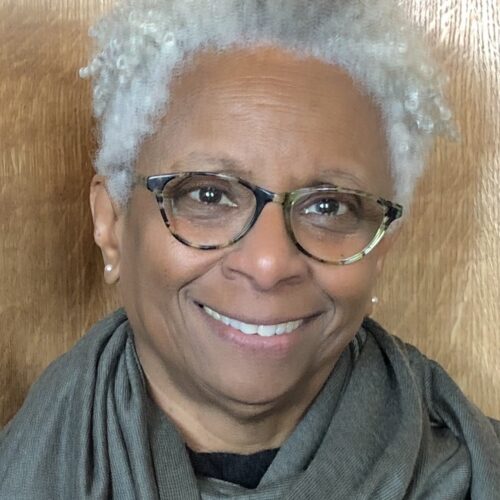 M. Shawn Copeland is smiling and wearing tortoise shell eyeglasses, pearl earrings, short white hair, with a gray-green scarf around her neck over a black shirt. The photo background is tan wood.